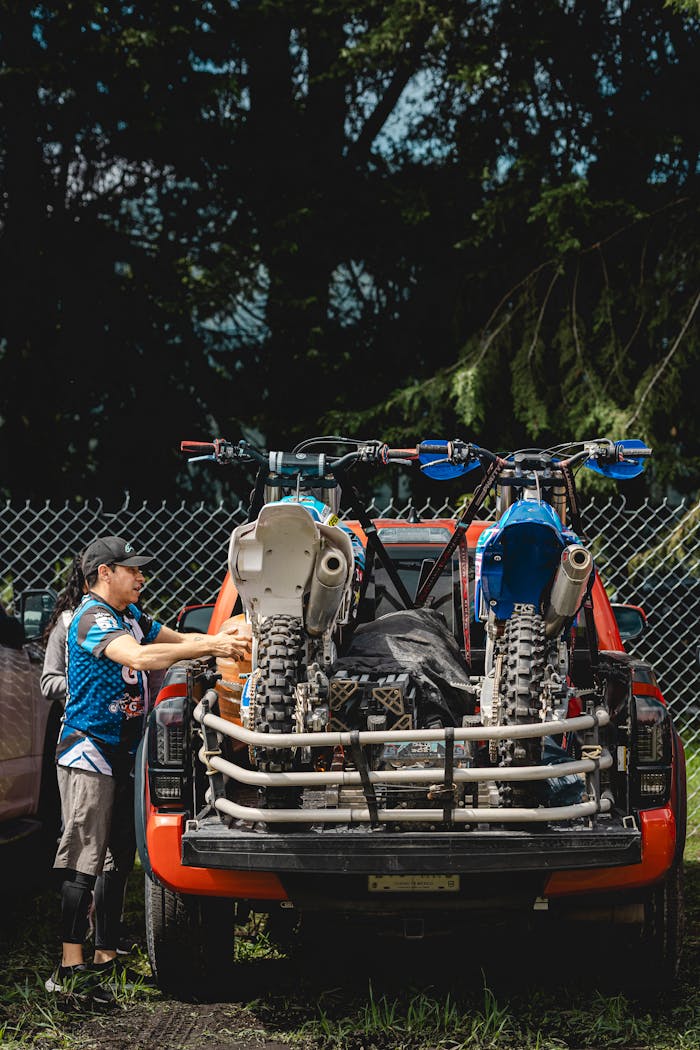 Two motocross bikes loaded on a pickup truck parked outdoors near a fence.